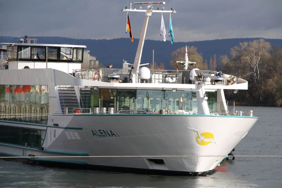 Ein modernes Flusskreuzfahrtschiff mit dem Namen ALENA, das am Ufer anlegt. Im Hintergrund sind Bäume und eine bewölkte Himmel sichtbar.