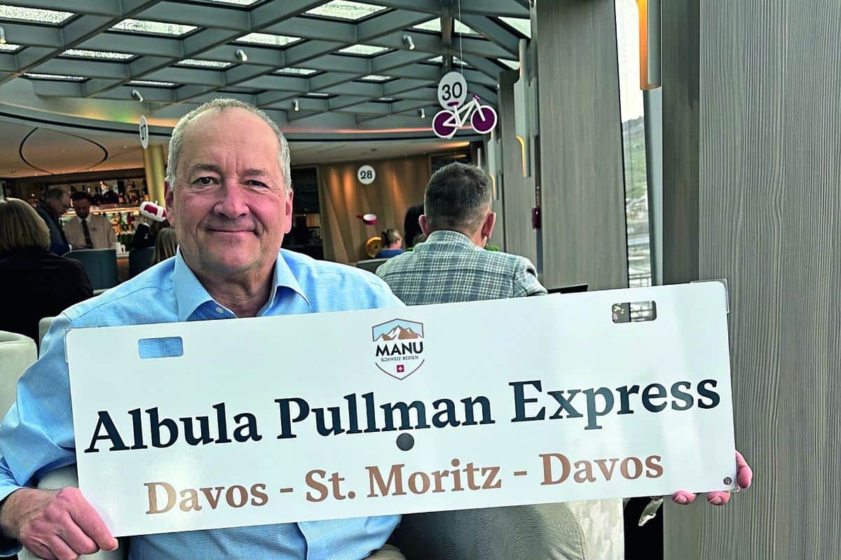 Ein Mann hält ein Schild mit der Aufschrift 'Albula Pullman Express' in einem modernen Innenraum mit anderen Gästen im Hintergrund.