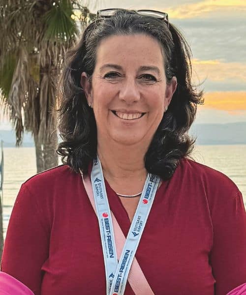 Eine lächelnde Frau mit schulterlangen, dunklen Haaren steht am Strand, umgeben von Palmen und einem Sonnenuntergang im Hintergrund.