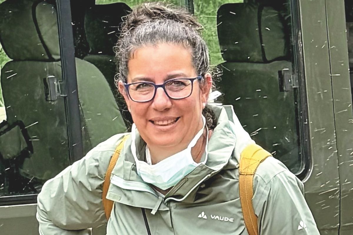 Eine Frau mit Brille und hochgesteckten Haaren trägt eine hellgrüne Vaude-Jacke und eine unter dem Kinn hängende medizinische Maske vor einem Geländewagen.