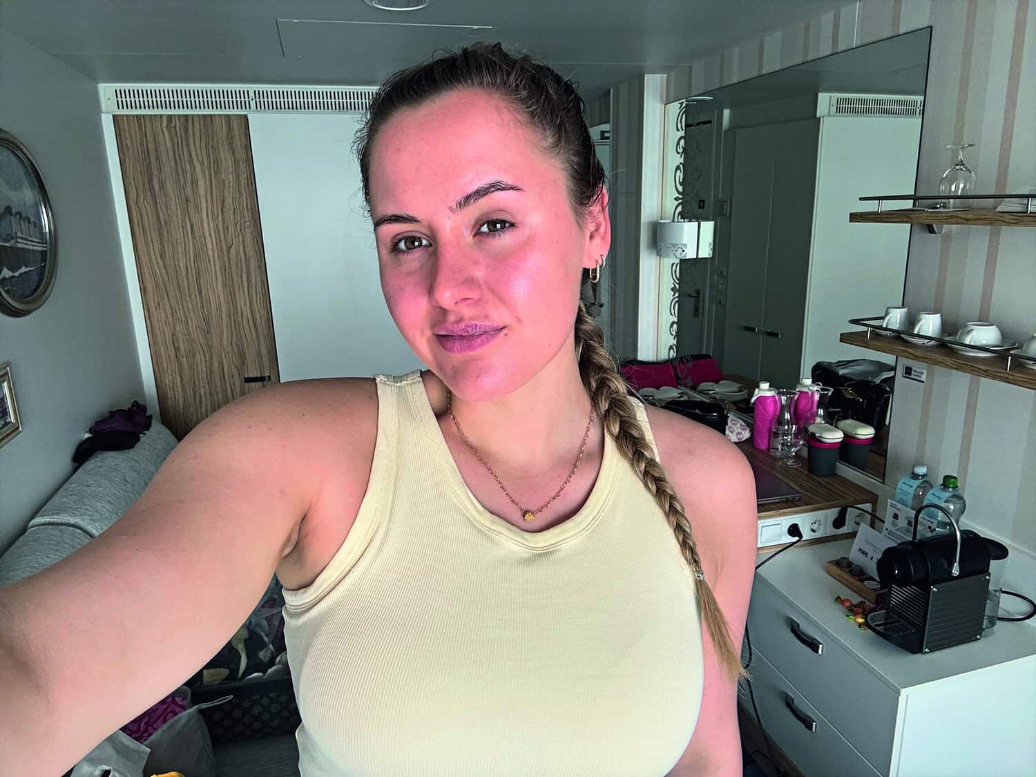 Eine junge Frau mit geflochtenem Haar steht in einem modernen Hotelzimmer vor einem Spiegel. Im Hintergrund sind Möbel und persönliche Gegenstände sichtbar.