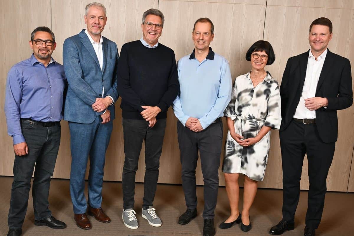 Sechs Personen stehen in einer Reihe vor einer Holzverkleidung, lächelnd und in formeller Kleidung, in einem modernen Innenraum.