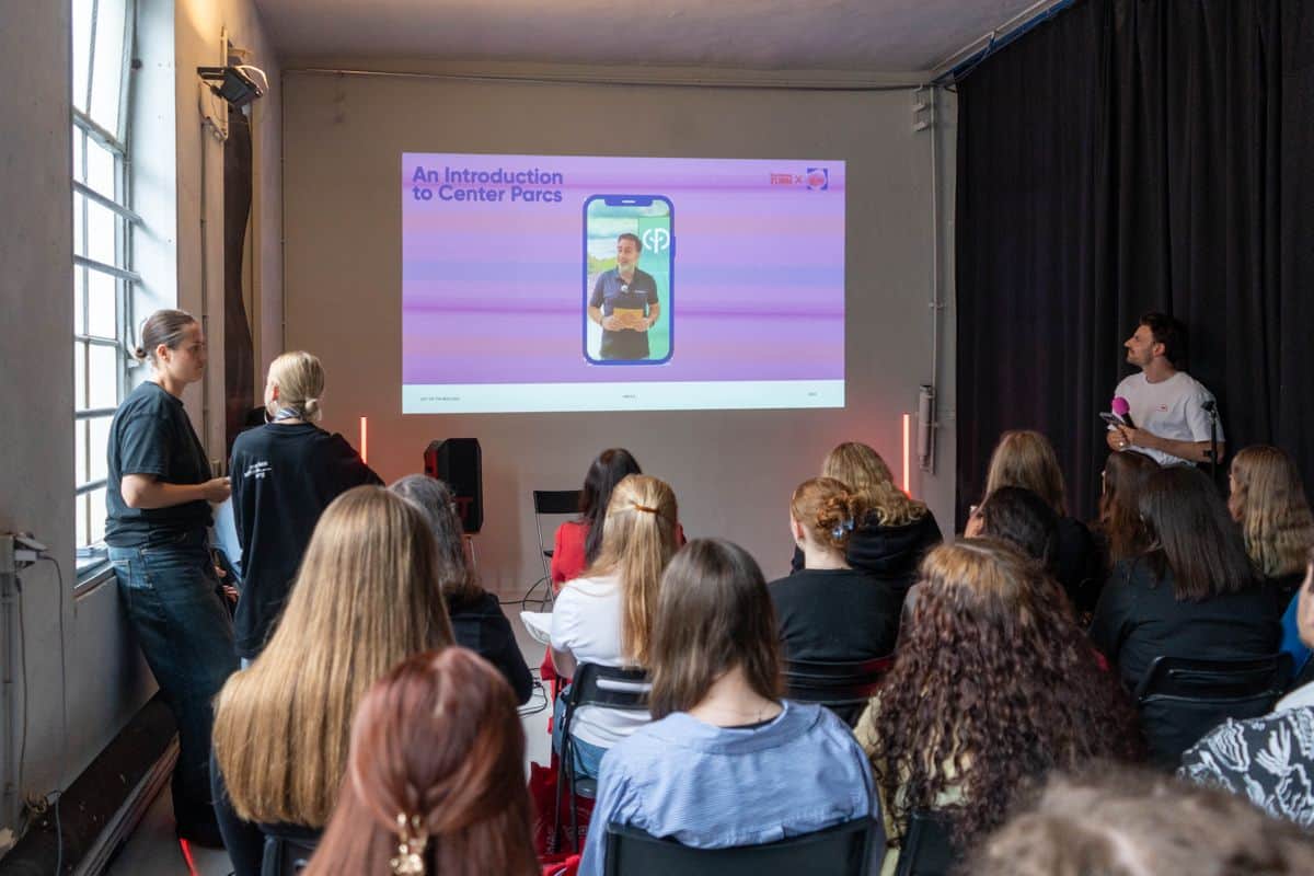 Eine Präsentation in einem Raum mit mehreren Zuhörern, die auf einen Bildschirm mit einer mobilen App-Demonstration schauen.