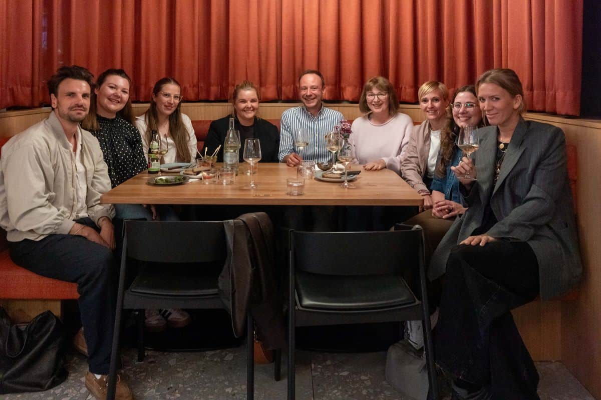 Eine Gruppe von neun Personen sitzt an einem Tisch in einem Restaurant mit orangefarbenen Vorhängen. Sie lächeln und halten Gläser in der Hand.