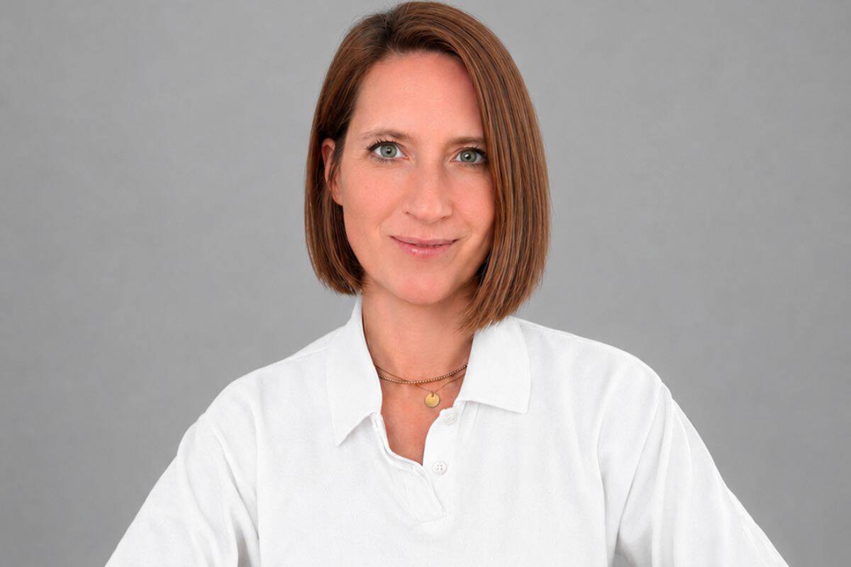 Porträt einer Frau mit schulterlangen braunen Haaren, die ein weißes Hemd trägt, vor einem grauen Hintergrund. Sie lächelt leicht und schaut direkt in die Kamera.