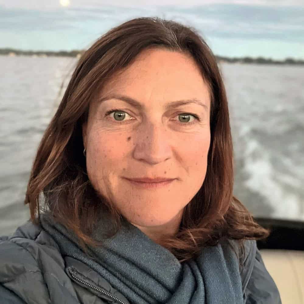 Porträt einer Frau mit braunen Haaren und grünen Augen, die auf einem Boot steht, im Hintergrund Wasser und Himmel sichtbar.