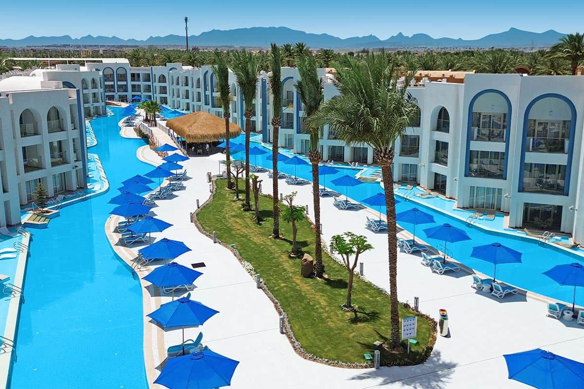 Luftaufnahme eines Resorts mit blauen Pools, Liegen und Sonnenschirmen, umgeben von Palmen und modernen Gebäuden im Hintergrund.