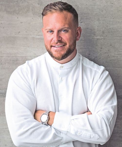 Ein Mann mit kurzem Bart und lächelndem Gesicht, gekleidet in ein weißes Hemd, steht vor einer grauen Betonwand mit verschränkten Armen.