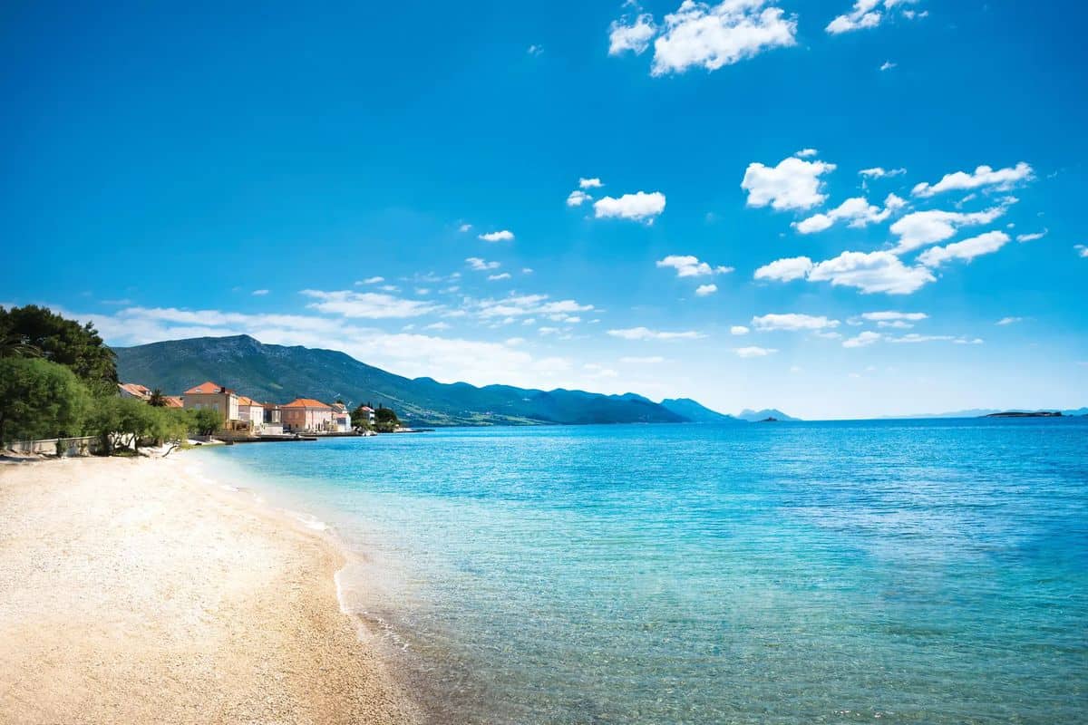 Ein klarer, blauer Himmel spiegelt sich im ruhigen Wasser einer Küste, umgeben von sanften Hügeln und einer kleinen Ansiedlung am Strand.