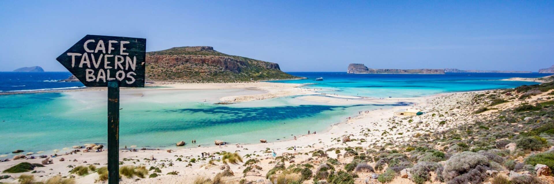 Ein Blick auf die Küste mit klarem, türkisfarbenem Wasser, einem Strand und einem Wegweiser zu einem Café und einer Taverne in Balos.
