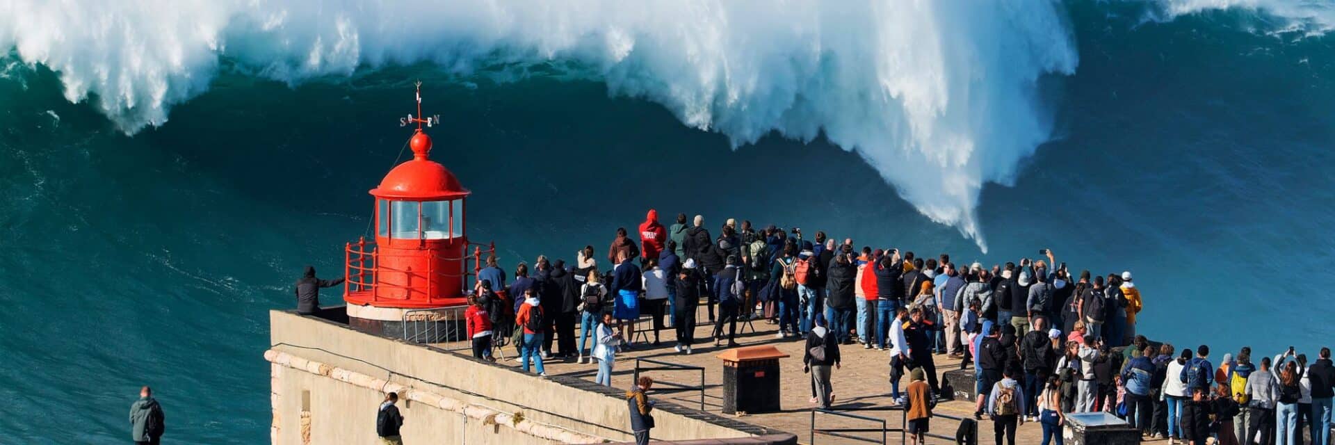 wm-portugal Eine große Menschenmenge steht auf einem Steg und beobachtet einen riesigen Wellenbrecher neben einem roten Leuchtturm.