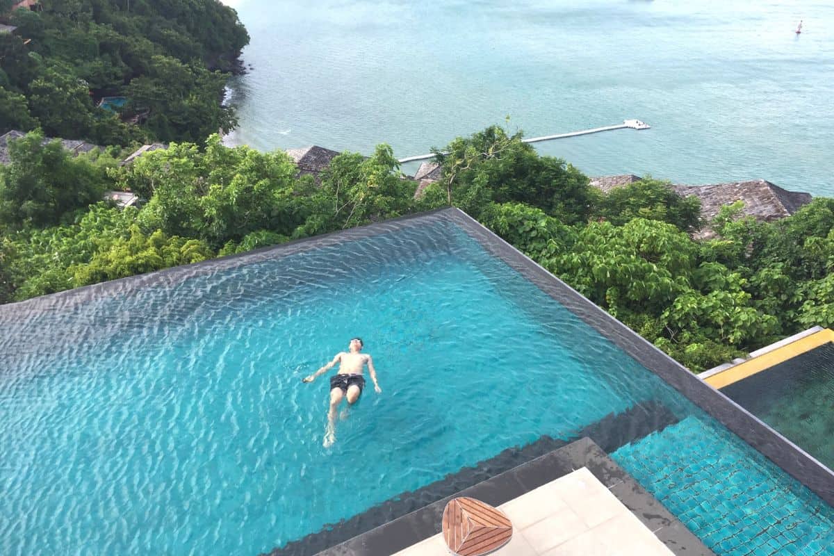 Ein Mann schwimmt entspannt in einem Infinity-Pool mit Blick auf das Meer und umgebende grüne Landschaft.
