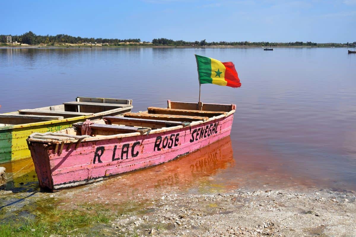 Bunte Fischerboote am Ufer des rosa gefärbten Lac Rose in Senegal, mit einer Flagge, die im Wind weht.
