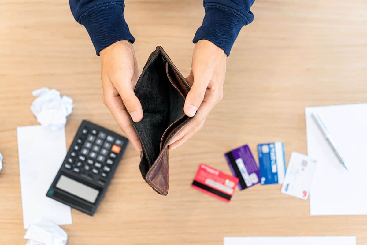 Eine Person hält eine leere Geldbörse in der Hand, umgeben von Kreditkarten, einem Taschenrechner und zerknülltem Papier auf einem Tisch.