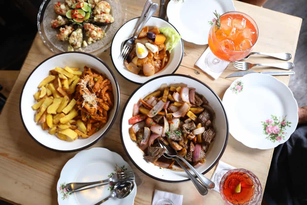 Ein Tisch mit verschiedenen Gerichten, darunter Pommes, Fleisch, Gemüse und ein Getränk in einem Glas, umgeben von Tellern und Besteck.