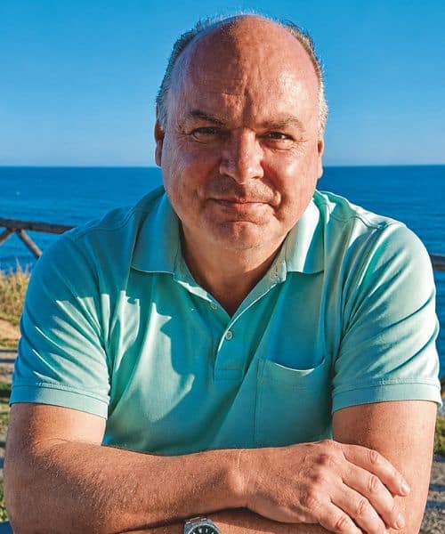 Ein Mann mit kurzem, grauem Haar sitzt am Strand, lächelt und blickt in die Kamera. Im Hintergrund ist das Meer und ein blauer Himmel zu sehen.
