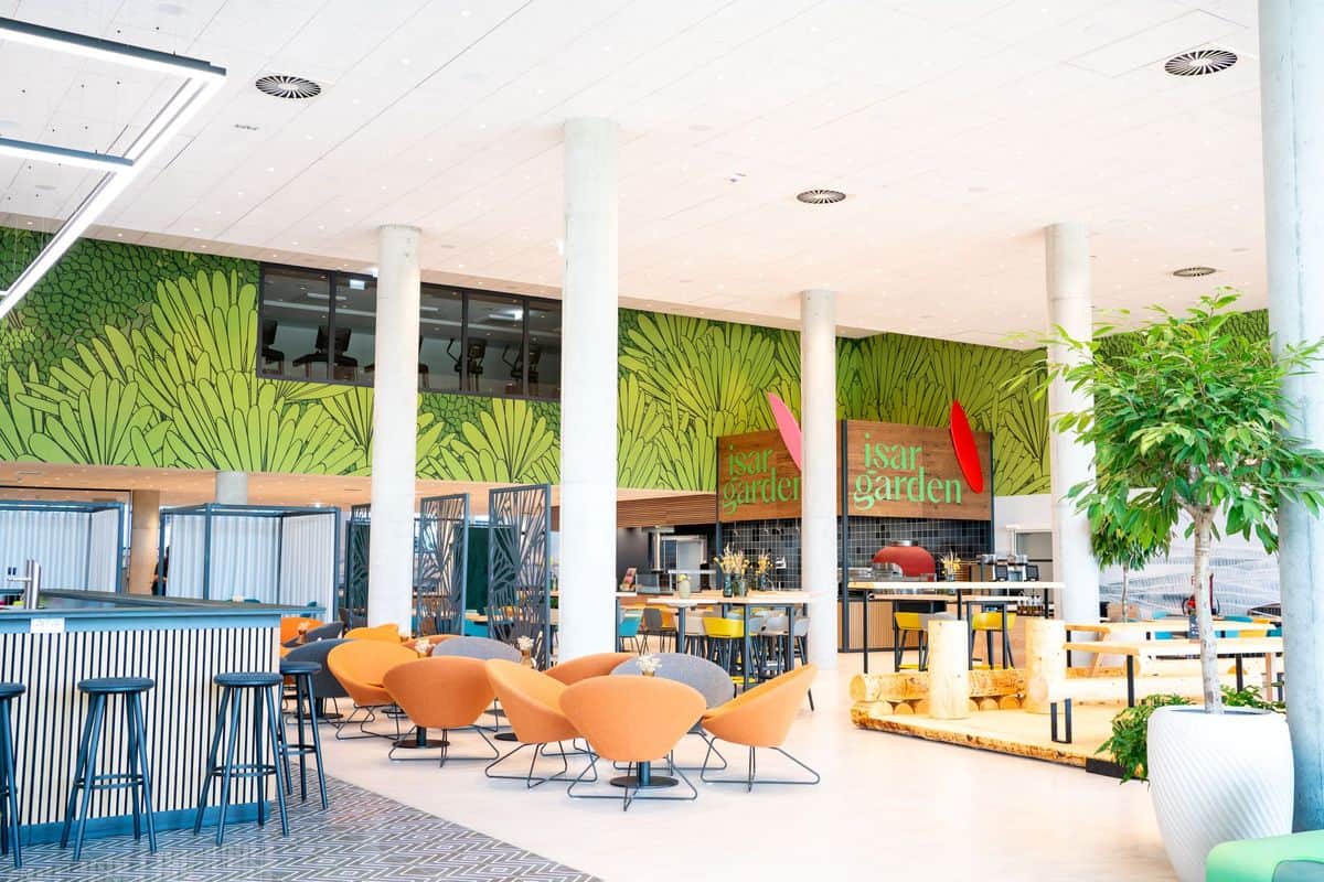Moderne Innenarchitektur eines Gastronomiebereichs mit bunten Stühlen, einem großen Wandgemälde und Pflanzen im Hintergrund.