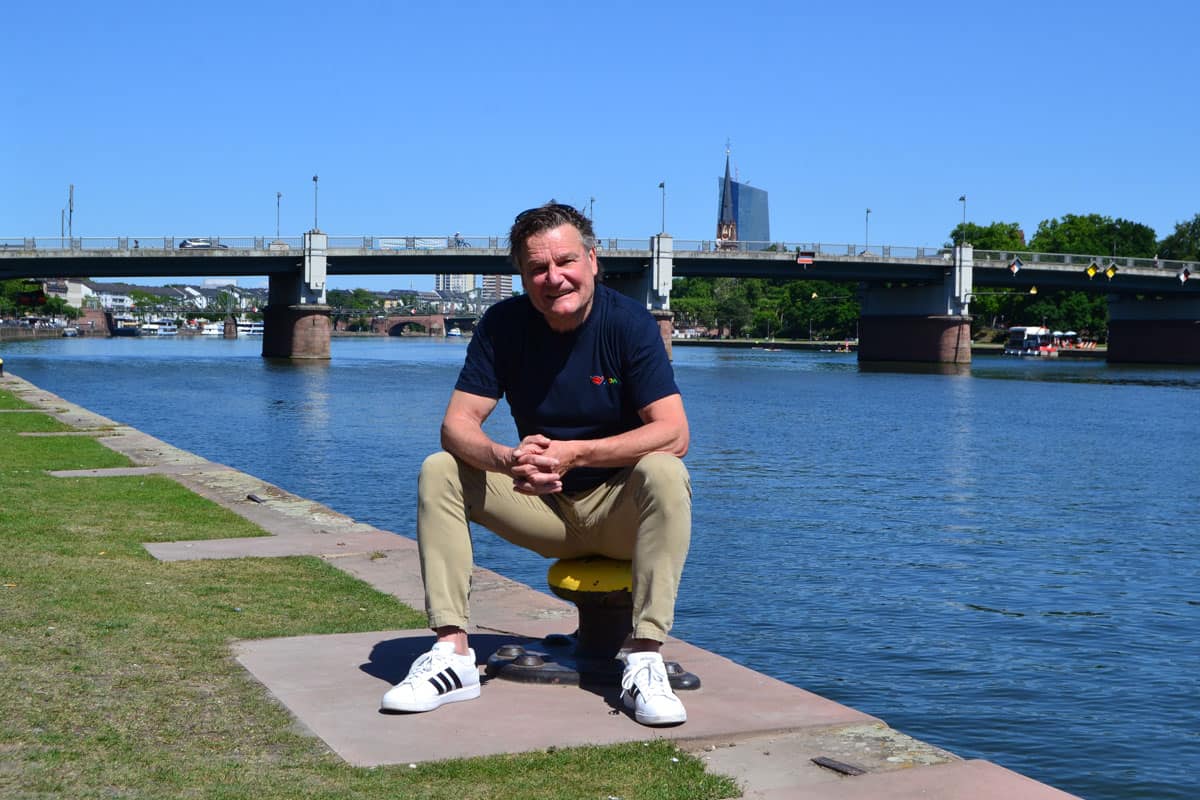 Ein Mann sitzt am Ufer eines Flusses, umgeben von grünem Gras, mit einer Brücke im Hintergrund und einem klaren blauen Himmel.
