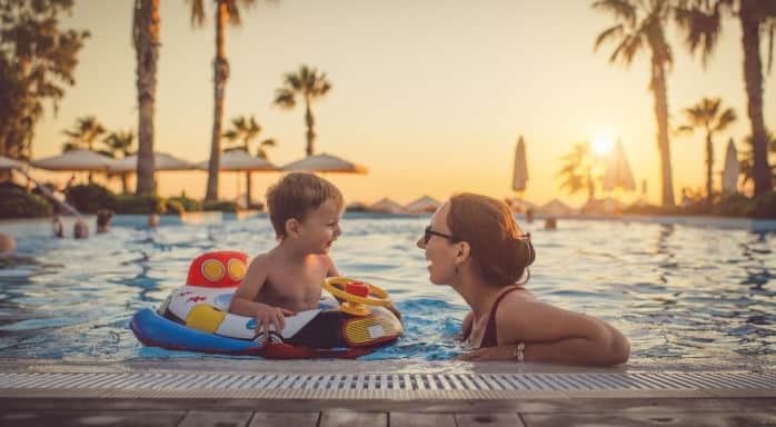 Eine Mutter und ihr kleiner Sohn im Pool, umgeben von Palmen und Sonnenliegen, während die Sonne am Horizont untergeht.