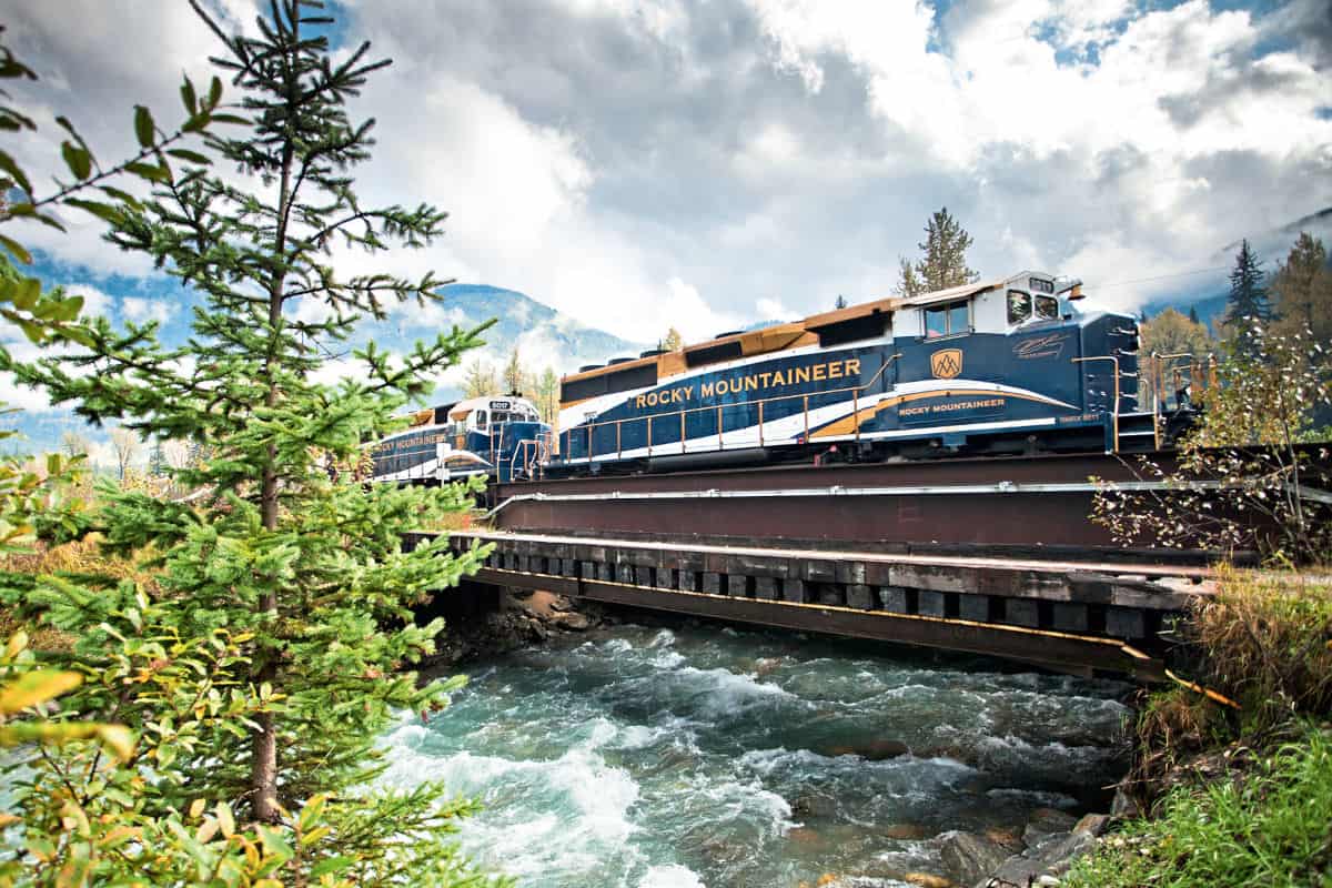 Ein Rocky Mountaineer Zug fährt über eine Brücke, umgeben von Bäumen und einem klaren Fluss unter einem bewölkten Himmel.