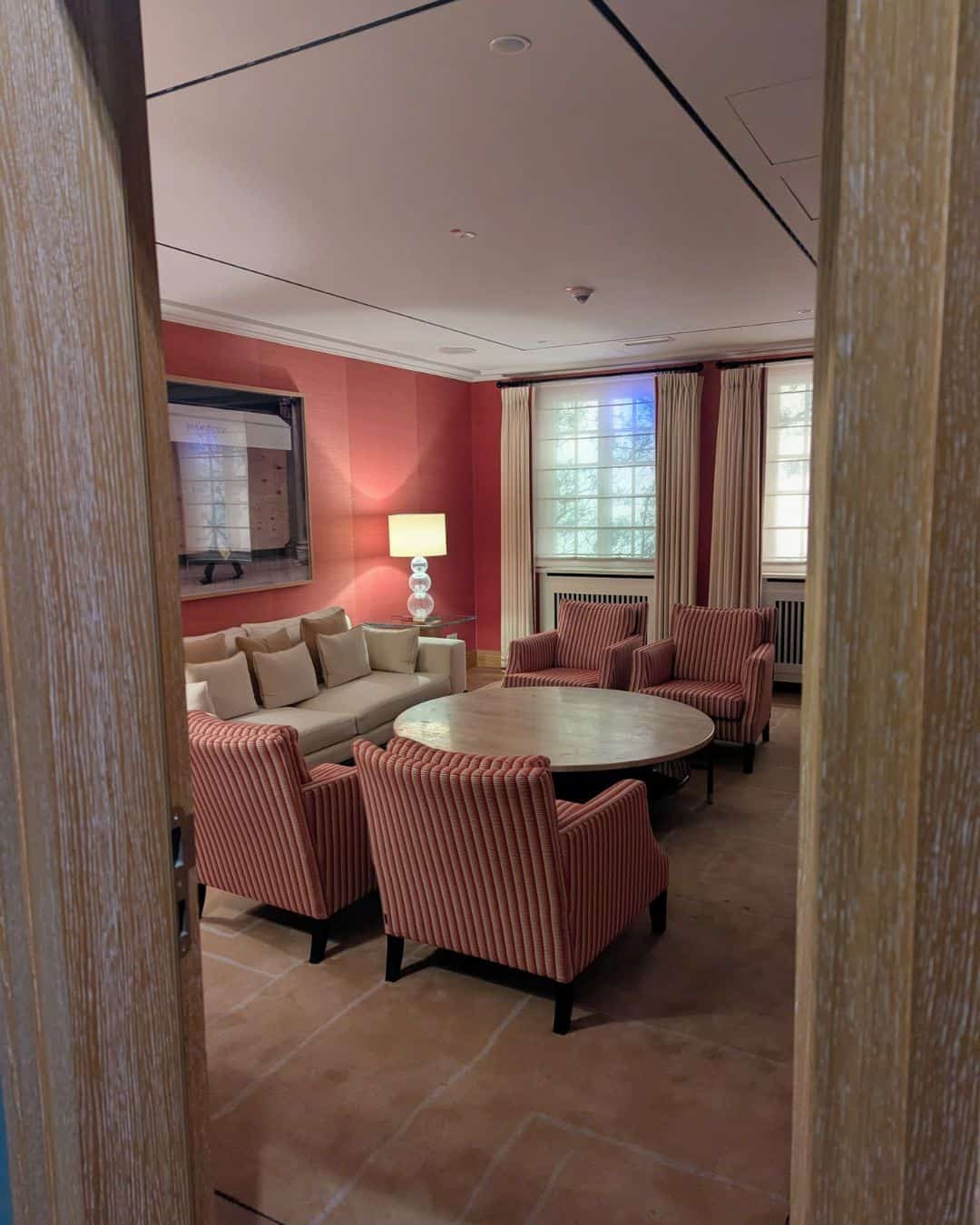 Ein eleganter Besprechungsraum mit einem runden Tisch, einem beigen Sofa und gestreiften Sesseln vor einer roten Wand und großen Fenstern.