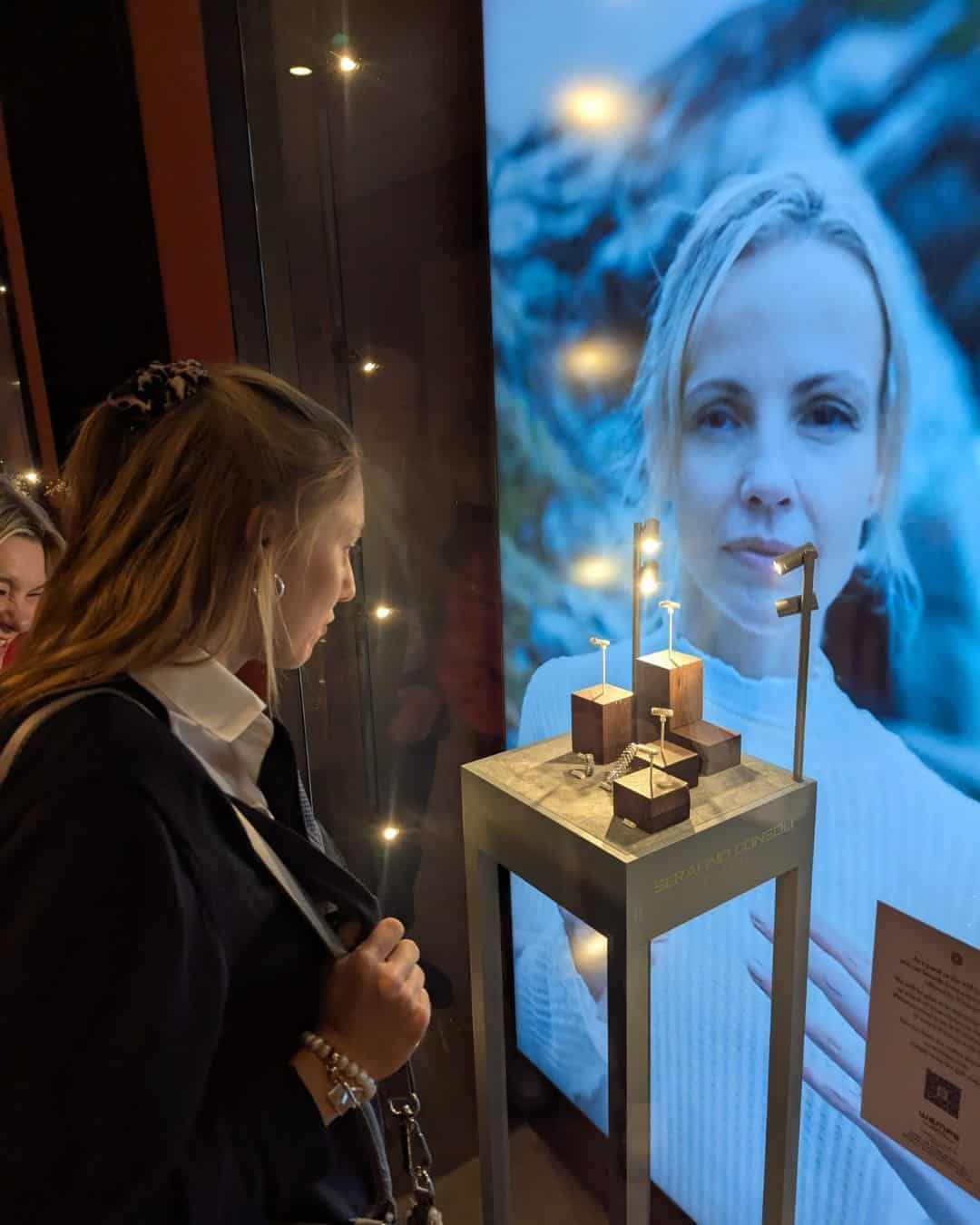 Eine Frau betrachtet eine Ausstellung mit Schmuckstücken auf einem Tisch, während ein großes Bild einer anderen Frau im Hintergrund zu sehen ist.