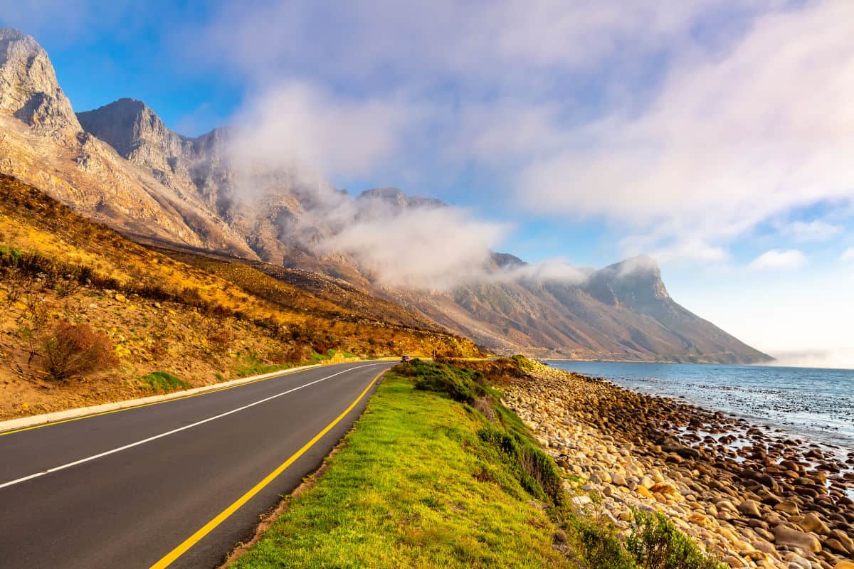 Eine kurvenreiche Straße verläuft entlang einer Küste mit felsigem Strand und majestätischen Bergen im Hintergrund, umhüllt von Nebel.