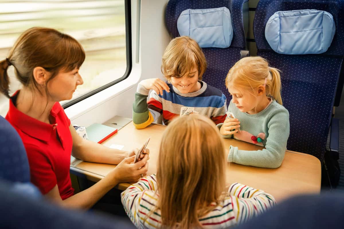 Eine Frau und drei Kinder sitzen an einem Tisch in einem Zugabteil und schauen auf ein Smartphone. Die Kinder sind aufmerksam und lächeln.