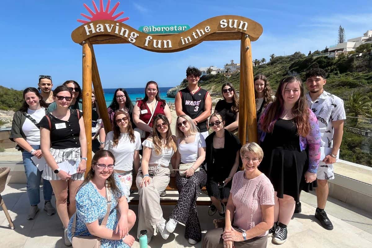 Gruppe von Personen posiert vor einem Holzrahmen mit der Aufschrift 'Having fun in the sun' und einem Blick auf das Meer im Hintergrund.