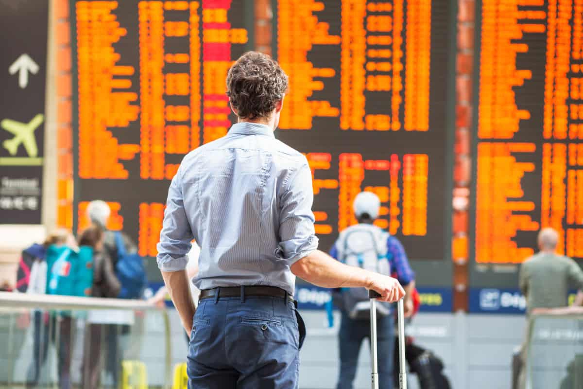 Ein Reisender mit einem Koffer steht vor einer großen Anzeigetafel am Flughafen, die Fluginformationen in leuchtenden Farben zeigt.