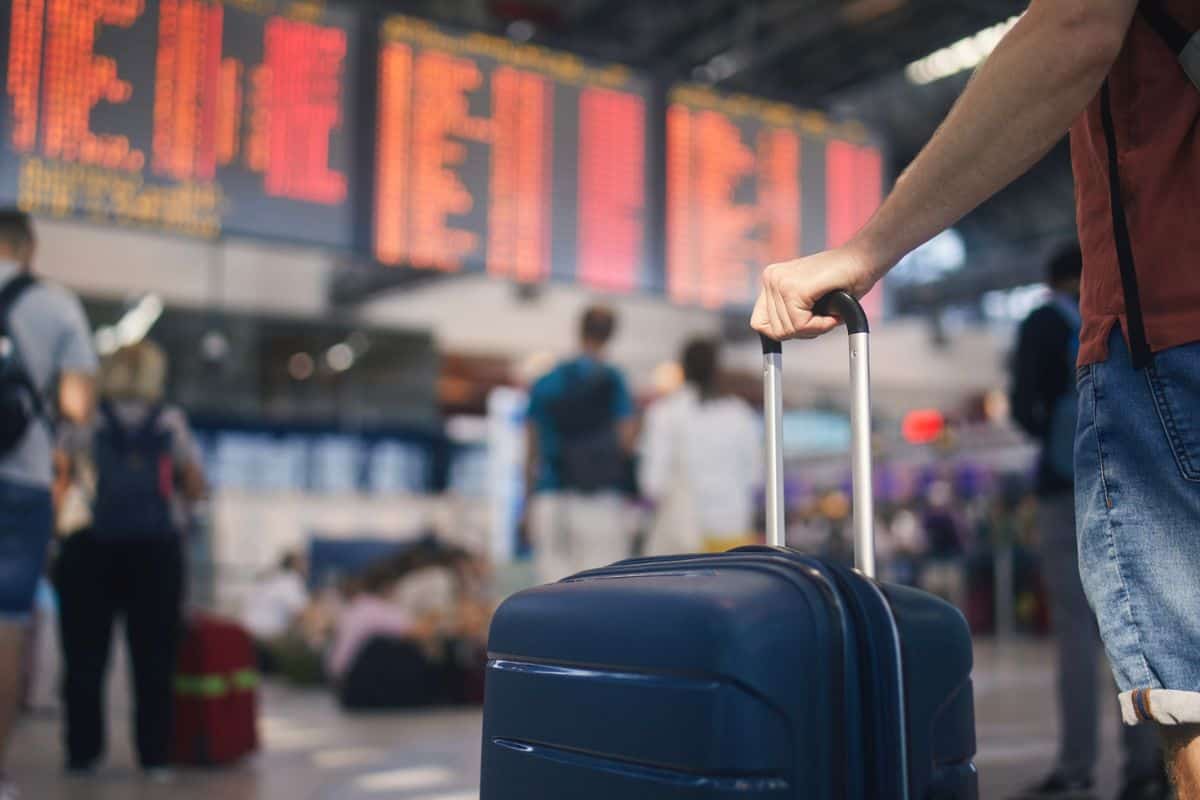 Ein Reisender hält einen Koffer in einem belebten Flughafen mit Anzeigetafeln im Hintergrund, die Fluginformationen anzeigen.