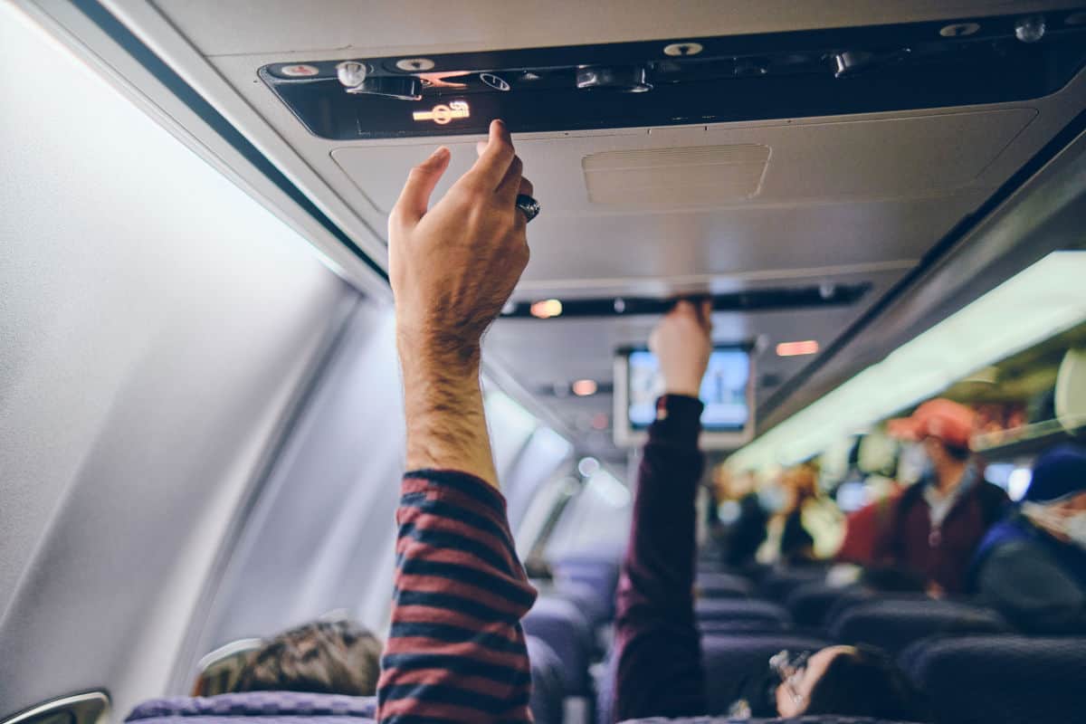 Zwei Hände heben sich in einem Flugzeug, um die Gepäckablage zu öffnen. Im Hintergrund sind weitere Passagiere und ein Bildschirm sichtbar.