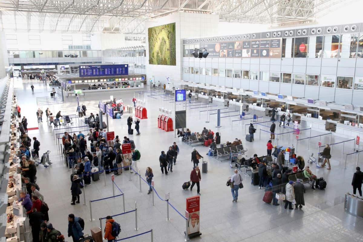 Blick auf einen modernen Flughafen mit Passagieren an Check-in-Schaltern und Wartenden in der Abflughalle. Helle, offene Architektur mit großen Fenstern.