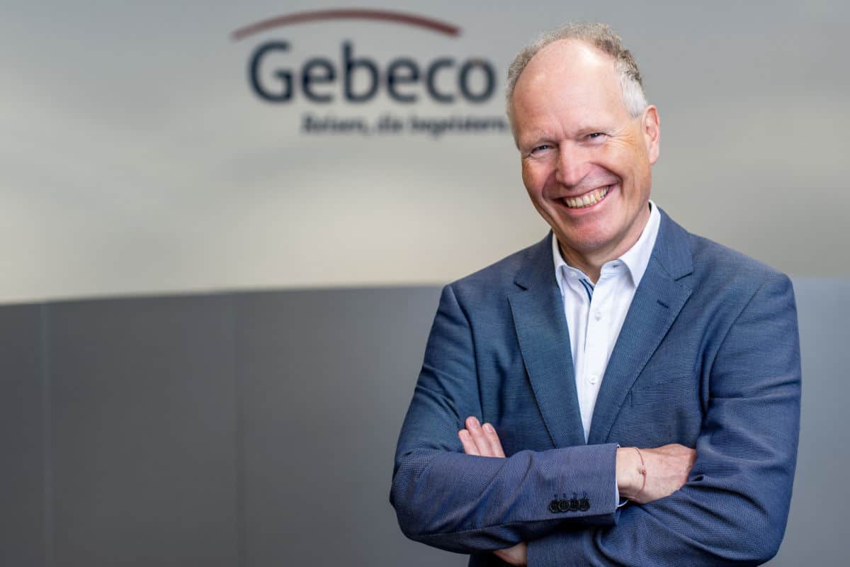Ein lächelnder Mann in einem blauen Anzug steht mit verschränkten Armen vor einer neutralen Wand mit dem Logo von Gebeco.