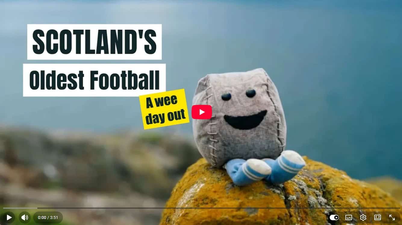 Ein lächelndes, plüschiges Fußballmaskottchen sitzt auf einem Felsen vor einem verschwommenen Hintergrund mit Wasser und Natur.