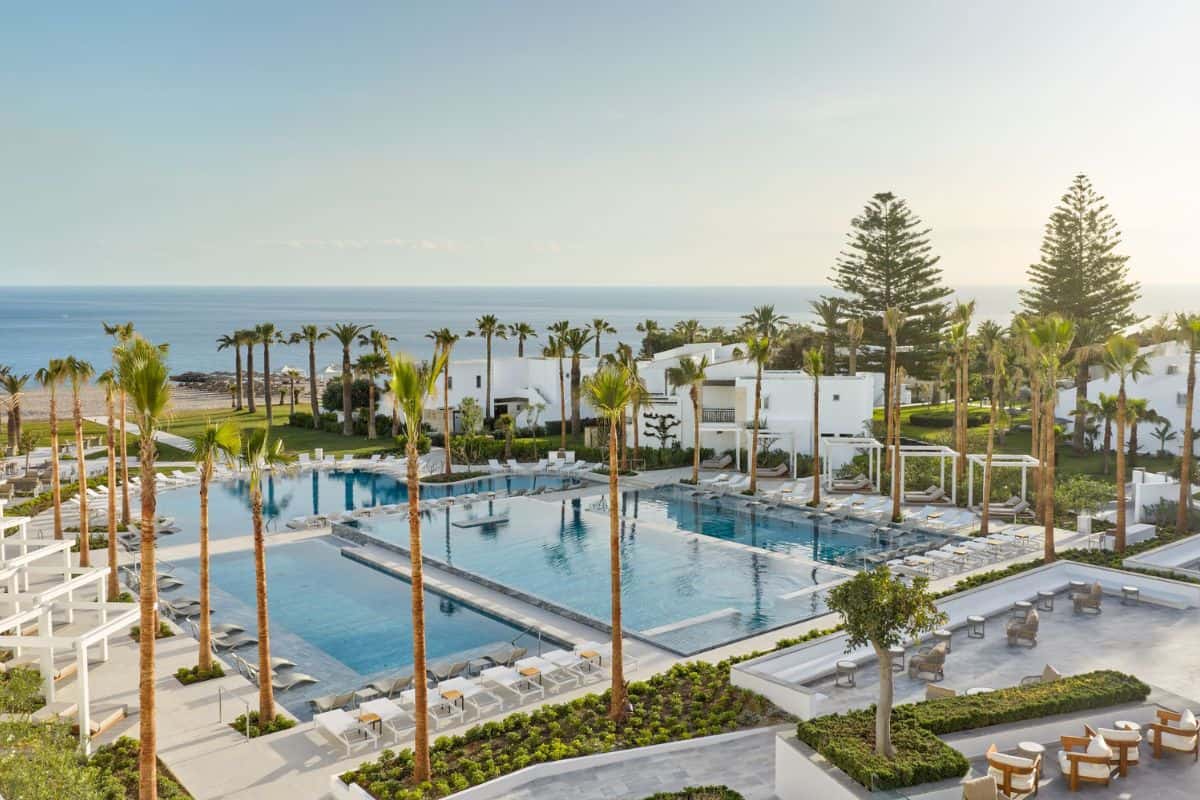 Blick auf einen modernen Poolbereich mit Palmen, Liegen und Meer im Hintergrund, umgeben von gepflegten Grünflächen und weißen Gebäuden.