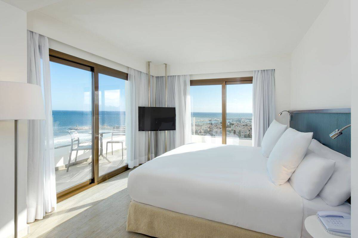 Helles Hotelzimmer mit Meerblick, großem Bett, Fernseher und Balkon. Große Fenster mit Vorhängen lassen viel Tageslicht herein.