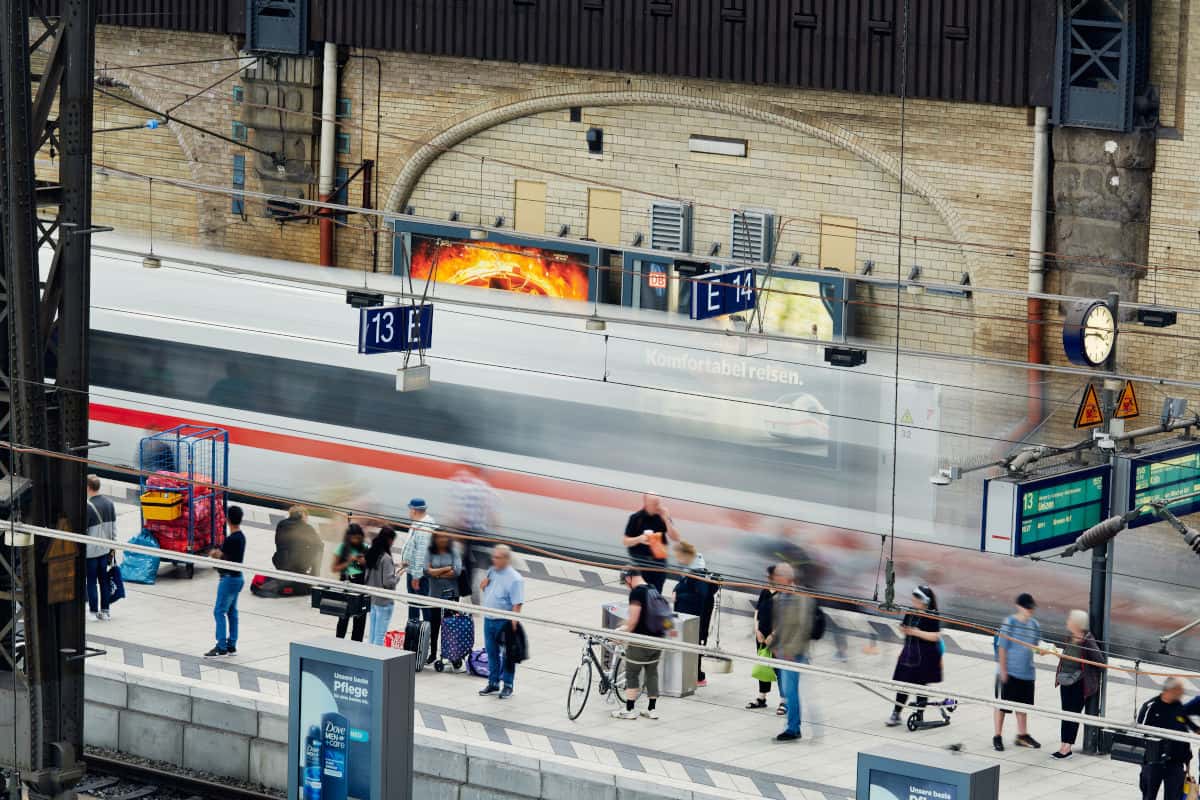 Der Hamburger Bahnhof mit Reisenden, einem Zug, der vorbeifährt, und Informationsanzeigen über Abfahrten.