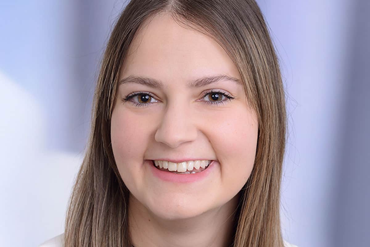 Porträt einer lächelnden Frau mit langen, glatten Haaren vor einem unscharfen, hellen Hintergrund.
