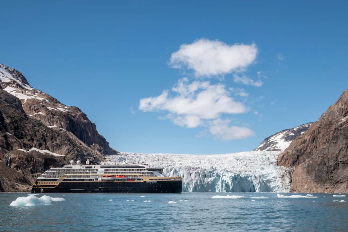 Ein Kreuzfahrtschiff liegt in einer ruhigen Bucht, umgeben von majestätischen Bergen und einem Gletscher unter einem klaren blauen Himmel.