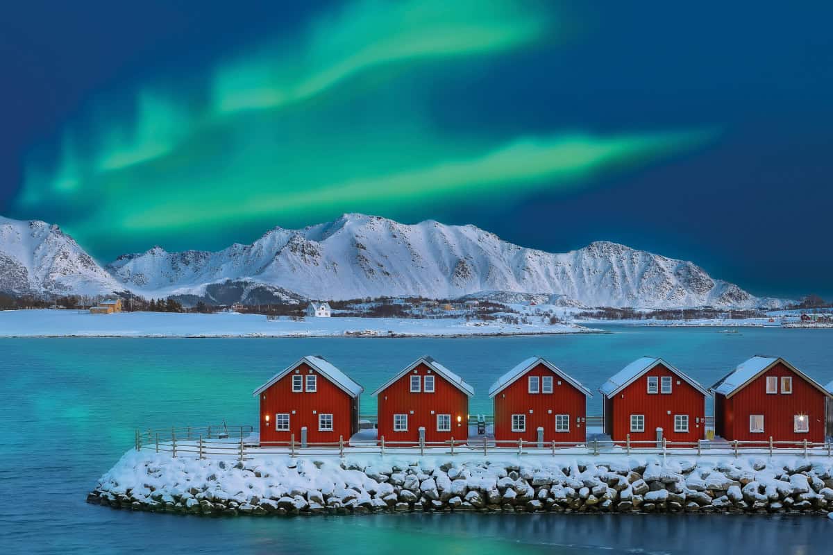 Rote Holzhäuser stehen auf einer schneebedeckten Insel, umgeben von einem klaren blauen Wasser und majestätischen Bergen unter einem grünen Polarlicht.