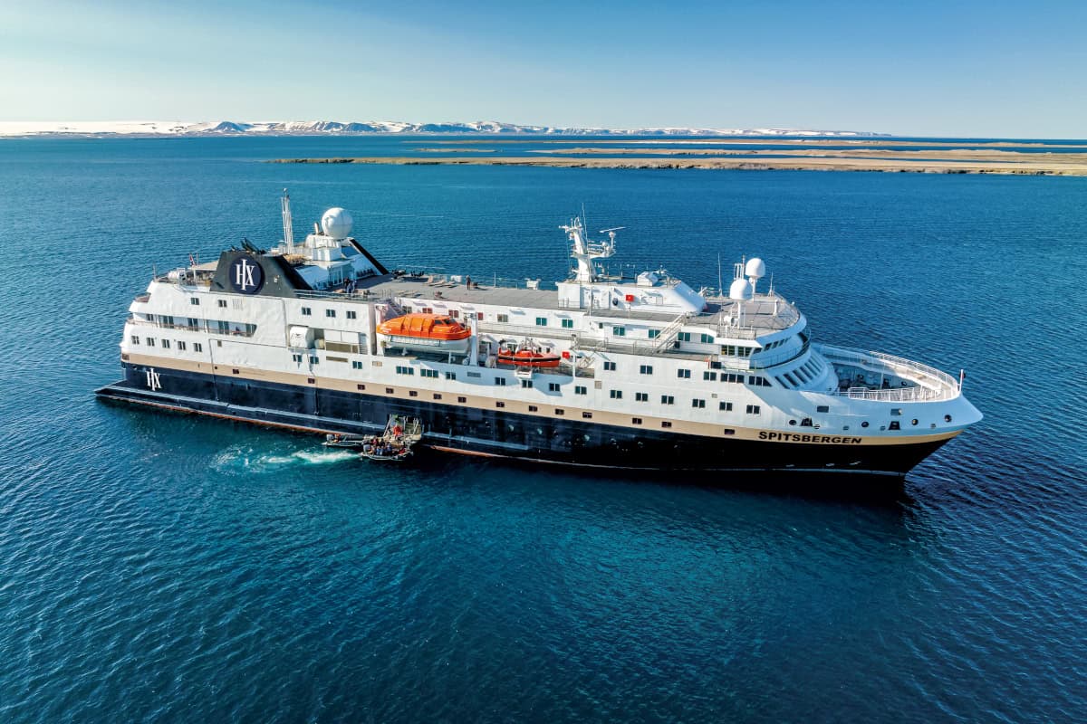 Ein Kreuzfahrtschiff mit dem Namen Spitsbergen fährt durch ruhiges, blaues Wasser, umgeben von einer malerischen Küstenlandschaft.