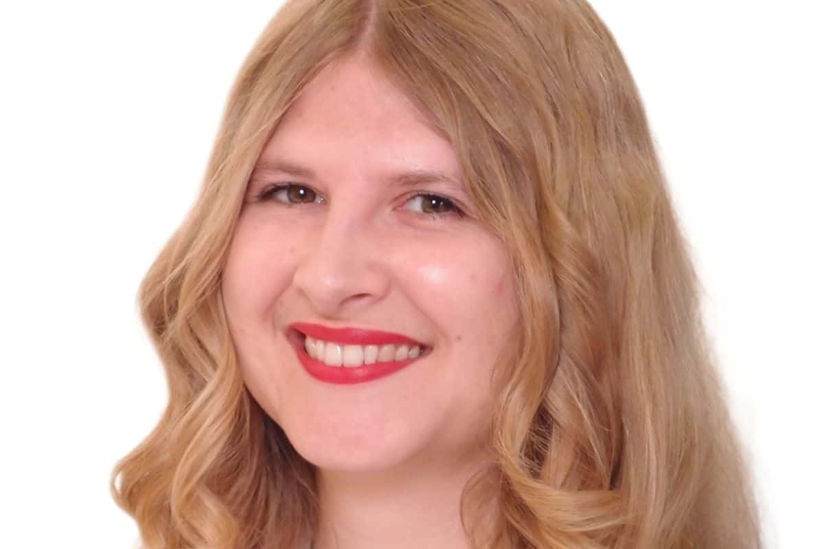 Porträt einer lächelnden Frau mit langen, lockigen, blonden Haaren und rotem Lippenstift vor einem weißen Hintergrund.