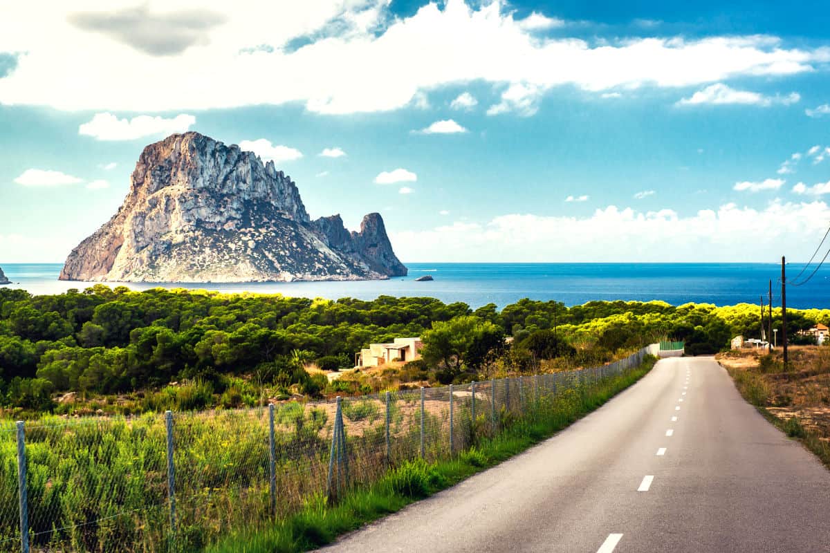 Panoramablick auf die Küste von Ibiza mit dem markanten Felsen am Strand Cala d'Hort und einer Straße, die durch die grüne Landschaft führt.