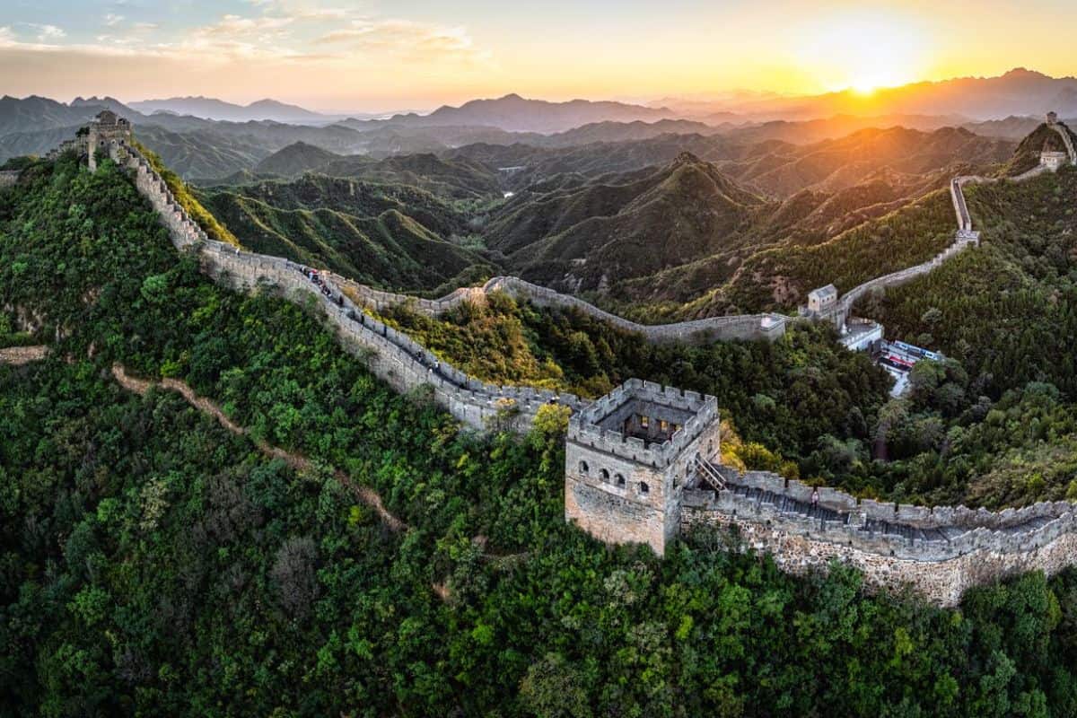 Luftaufnahme der Chinesischen Mauer, die sich über grüne Hügel erstreckt, mit Sonnenuntergang im Hintergrund und sanften Bergketten.