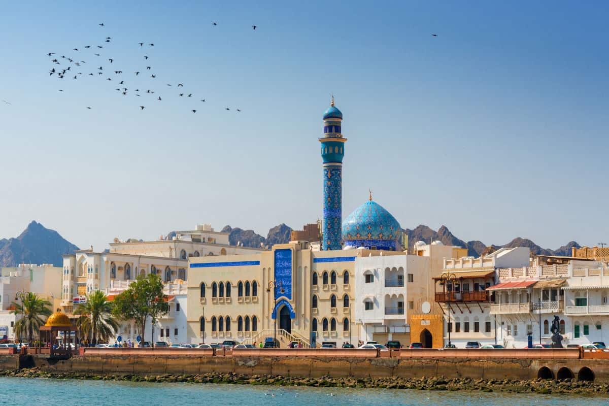 Blick auf eine Küstenstadt mit traditioneller Architektur, einer blauen Kuppelmoschee und Bergen im Hintergrund unter klarem Himmel.