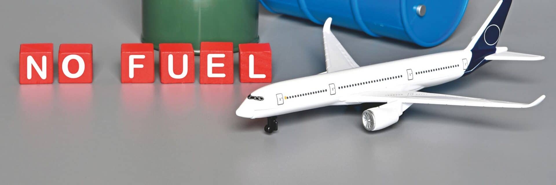 Ein Modellflugzeug steht neben roten Buchstaben, die 'NO FUEL' bilden, auf einer grauen Oberfläche mit einem grünen und blauen Fass im Hintergrund.