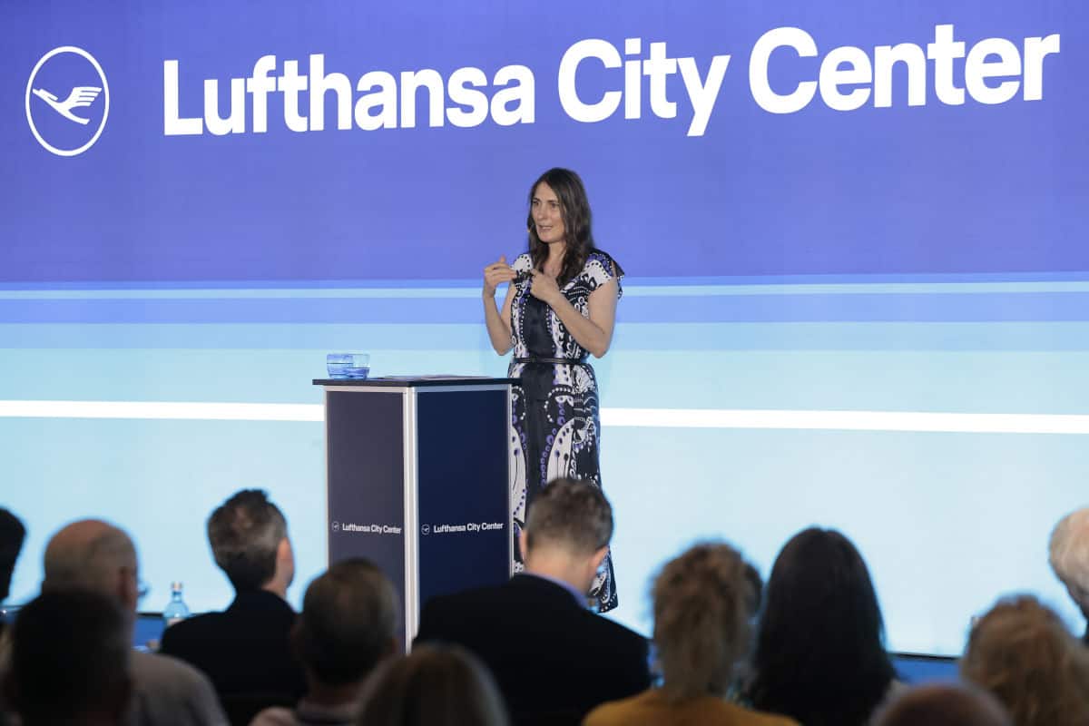 Eine Frau in einem schwarz-weißen Kleid steht an einem Podium vor einer großen Präsentationsleinwand mit dem Logo des Lufthansa City Centers.