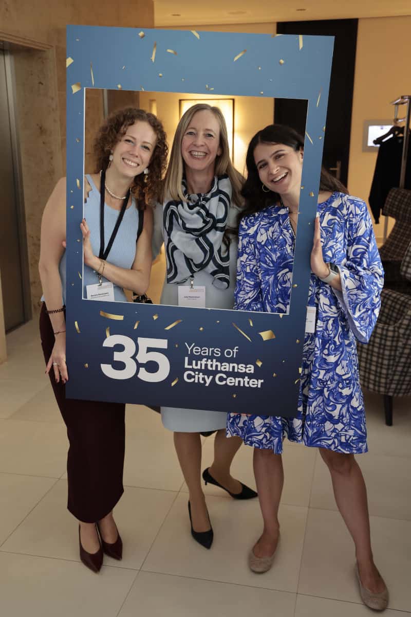 Drei Frauen posieren fröhlich vor einem dekorativen Rahmen mit der Aufschrift '35 Years of Lufthansa City Center' in einem modernen Innenraum.