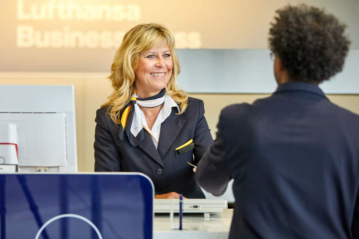 Eine Lufthansa-Mitarbeiterin am Check-in-Schalter lächelt freundlich, während sie einen Passagier am Counter bedient.